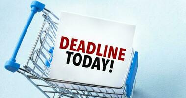 Shopping cart and text DEADLINE TODAY on white paper note list. Shopping list concept on blue background. photo