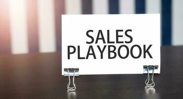 SALES PLAYBOOK sign on paper on dark desk in sunlight. Blue and white background photo