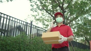 A parcel delivery worker wearing a red uniform is holding a parcel box to the recipient. At that time, he was using a mobile phone. contact the receiver in front of the house video