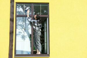 un mujer a mano lavados el ventana de el casa con un trapo con un rociar limpiador y un fregona afuera. la seguridad a altura, restaurar orden y limpieza en el primavera, limpieza Servicio foto