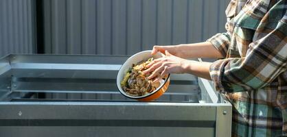 un mujer en un tartán camisa vierte comida residuos desde un cuenco dentro un compost montón de patata y Zanahoria peladuras. compost caja hecho de metal, Respetuoso del medio ambiente fertilizante para el jardín foto