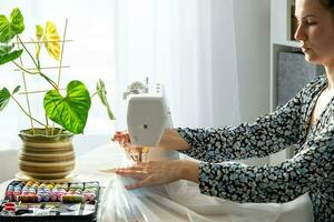 un mujer cose tul en un eléctrico de coser máquina en un blanco moderno interior de un casa con grande ventanas, casa plantas. comodidad en el casa, un ama de casa pasatiempo foto