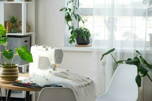 Sewing machine on a round table in the white interior of the house near the window with a transparent curtain, and a home plant in a pot - a modern interior. hobby, home business photo