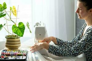 A woman sews tulle on an electric sewing machine in a white modern interior of a house with large windows, house plants. Comfort in the house, a housewife's hobby photo