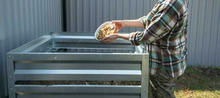 A woman in a plaid shirt pours food waste from a bowl into a compost heap of potato and carrot peelings. Compost box made of metal, eco-friendly fertilizer for the garden photo