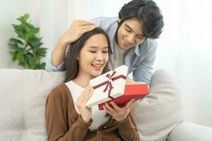 Celebrating anniversary, surprise. Relationship asian young couple love, boyfriend use hand cover girlfriend eye, give a gift by hide box at behind, woman getting present while sitting on sofa at home photo