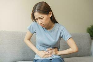 Flatulence asian young woman hand in stomach ache, suffer from food poisoning, abdominal pain and colon problem, gastritis or diarrhoea sitting on sofa at home. Patient belly, abdomen or inflammation. photo