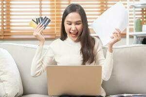 Happy excited, asian young woman, girl holding credit card and paperwork getting, received job promotion, approve tax refund cash back, looking good news at laptop computer, sitting on sofa at home. photo