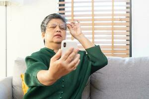 Presbyopia, Hyperopia mature asian woman holding eyeglasses having problem with vision problem trying to read text on mobile, eye disease of old elderly sitting on couch. Poor eyesight, health care. photo