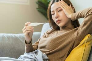 enfermo, influenza asiático joven mujer, niña dolor de cabeza tener fiebre, mano conmovedor frente, participación termómetro para cheque medida cuerpo temperatura, enfermedad mientras sentado descanso en sofá a hogar. salud cuidado. foto