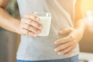 dolor, alergia joven asiático mujer, niña mano en barriga sufrir después bebida vaso de leche, infeliz y malo estómago dolor. lactosa intolerancia y lechería comida , salud problema concepto. foto