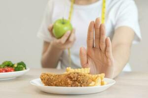 dieta, dieta asiático joven mujer mano en empujar afuera, negar frito pollo, francés frito y escoger verde manzana, vegetales ensalada, comer comida para bueno saludable, salud cuando hambriento. hembra peso pérdida gente. foto