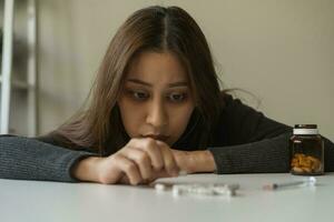 Anti drugs, drug addict asian young woman hand holding pills, medicament with narcotic syringe on table at home, abuse overdose. Sick pain of health, unhealthy people. Suicide depressed or despair. photo