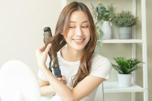 hermosa Rizado morena, atractivo asiático joven mujer, niña sonriente utilizando bigudí, curling hierro largo pelo con cerámico bigudí plancha de pelo a hogar. peinado y peluquería. belleza y pelo cuidado . foto