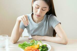 dieta en aburrido rostro, infeliz hermosa asiático joven mujer, niña en haciendo dieta, participación tenedor en ensalada lámina, disgusto o cansado con comer Fresco vegetales. nutrición de limpio, sano comida bueno gusto. foto