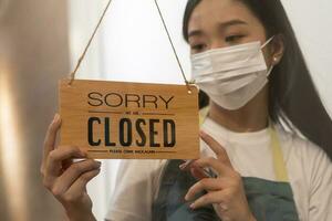 Asian young owner retail,coffee shop woman turning sign board to closed with wearing face mask,protection to pandemic of coronavirus.Close store,restaurant or pub due to lockdown, quarantine of covid. photo