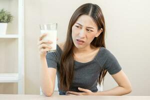 dolor, alergia asiático joven mujer, niña mano en barriga sufrir después bebida vaso de leche, sufrir y malo estómago dolor. lactosa intolerancia y lechería comida , salud problema concepto. foto