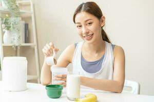 dieta comida reemplazo para peso pérdida, sonrisa asiático joven mujer, niña en ropa de deporte, mano en participación cucharón haciendo proteína agitar, bebida suplemento para músculo después rutina de ejercicio a hogar. sano cuerpo cuidado. foto