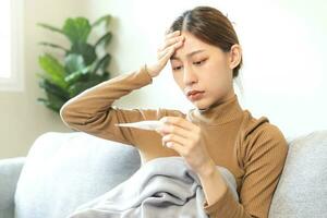 Sick, influenza asian young woman, girl headache have fever, hand touching forehead, holding thermometer for check measure body temperature, illness while sitting rest on sofa at home. Health care. photo