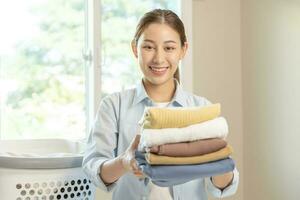 ama de casa, atractivo asiático joven mujer, niña participación muchos plegable ropa en el cesta después Lavado limpiar ropa y el secado, casa trabajando en habitación a hogar, mucama faena lavandería ordenado. foto