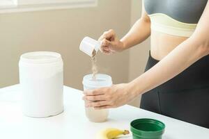 dieta comida reemplazo para peso pérdida, sonrisa asiático joven mujer, niña en ropa de deporte, mano en participación cucharón haciendo proteína agitar, bebida suplemento para músculo después rutina de ejercicio a hogar. sano cuerpo cuidado. foto