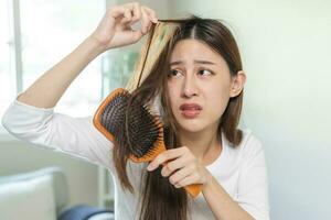 grave, preocupación asiático joven mujer, niña participación cepillar, espectáculo su peine, cepillo para el pelo con largo pérdida pelo problema después cepillado, pelo otoño fuera en su mano en vivo habitación. salud cuidado, belleza tratamiento. foto