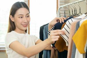 elección de ropa, nada a tener puesto. atractivo asiático joven mujer, niña tratar en aparecer, contento elegir vestido, atuendo en percha en guardarropa en habitación armario a hogar. decidiendo blusa qué a poner en cuales uno foto