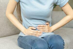 Flatulence asian young woman hand in stomach ache, suffer from food poisoning, abdominal pain and colon problem, gastritis or diarrhoea sitting on sofa at home. Patient belly, abdomen or inflammation. photo