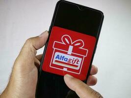 Jakarta, Indonesia, 2023. Hand holding mobile phone with finger touch the Alfa Gift logo on screen. photo