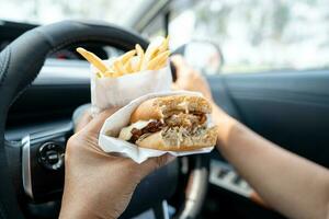 asiático dama participación hamburguesa y francés papas fritas a comer en auto, peligroso y riesgo un accidente. foto