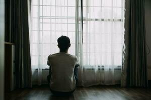 solo hombre silueta curioso a el ventana cerrado con cortinas en dormitorio. hombre soportes a ventana solo foto