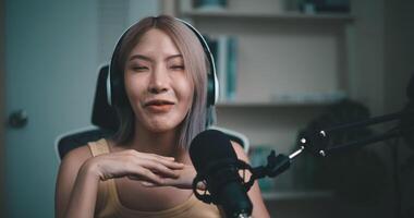 Footage of Young Asian woman influencer wearing headphones talking into a microphone while recording a radio show in a living room home studio. Content creator and influencer marketing concepts. video