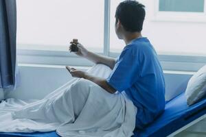 Asian male patient lying on bed with face mask in recovery room in hospital ward. All people wearing mask to prevent covid19 virus infection during coronavirus pandemic. The man feels lonely and bore. photo