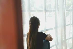 Thoughtful girl sitting on sill embracing knees looking at window, sad depressed teenager spending time alone at home, young upset pensive woman feeling lonely or frustrated thinking about problems photo