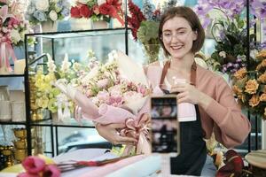 E-commerce business, young White female florist influencer demonstrates and shows floral arrangements, online live streaming with smartphone application in flower shop, beautiful blossoms store. photo