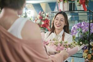 joven asiático hembra florista trabajador en delantal entrega hermosa Fresco flores ramo de flores a cliente quien comprado orden, contento vendedor quien trabajos en vistoso flor comercio, y pequeño negocio emprendedor. foto