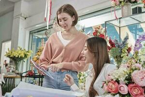 Two young beautiful female florist partners discuss bloom bouquet arrangement design in tablet, online purchase order work in colorful flower shop store with fresh flora, SME business entrepreneur. photo