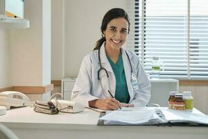 retrato de hermosa hembra médico y profesional nutricionista en uniforme sonriente y mirando a cámara con suplementario comidas en escritorio para sano dieta a hospital. foto