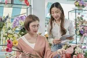 Two young beautiful female florist partners discuss bloom bouquet arrangement design in tablet, online purchase order work in colorful flower shop store with fresh flora, SME business entrepreneur. photo