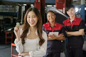 Young Asian female customer shows car key with automotive mechanic workers, happy smile in quality trust at maintenance garage, professional vehicle service center, fix check and repair auto industry. photo