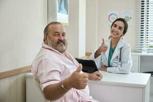 hermosa hembra médico en uniforme y blanco masculino paciente sonriente y mirando a cámara, salud chequeo cita a un trabajando escritorio, sano dieta clínica hospital. foto