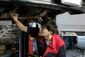 Professional young Asian male motor mechanic inspects undercarriage of electric car lifted by forklift jack for repair at garage, automotive maintenance service works industry occupation business. photo