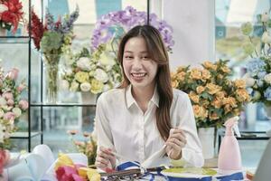 One Young beautiful Asian female florist entrepreneur arranging a bunch of blossoms, decorating with lovely ribbons, happy work in colorful flower shop store with blooms, and small business owner. photo