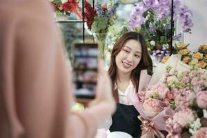 E-commerce business, young Asian female florist worker demonstrates and shows floral arrangements via online live streaming by smartphone application in bright flower shop, beautiful blossoms store. photo