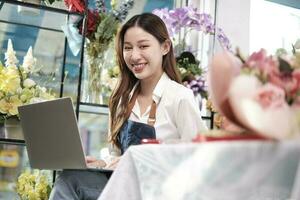 One young Asian female florist and SME entrepreneur in apron, works with laptop, online selling floral arrange in colorful flower shop store with beautiful bunch of blossoms, and e-commerce business. photo