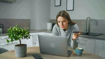 Frau Stehen im Zuhause gemütlich Küche, gelehnt Über das Tabelle und macht ein online Einkaufen mit ein Anerkennung Karte und Laptop video