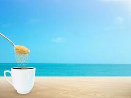 Spoon pouring granulated sugar into coffee mug on wooden table with the sea in the background photo