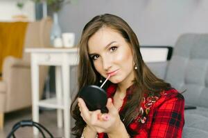 Young Caucasian woman in a red plaid shirt and skirt does her makeup, paints her lips with glitter at home, sitting on the couch and looking in the mirror photo