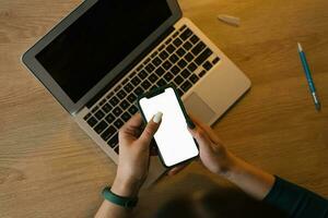 Mockup of the smartphone screen in the hands of a young woman against the background of a laptop at the table photo