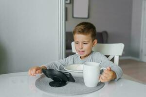 Child, a boy of five years old, is having breakfast at the kitchen table and watching cartoons on his smartphone photo